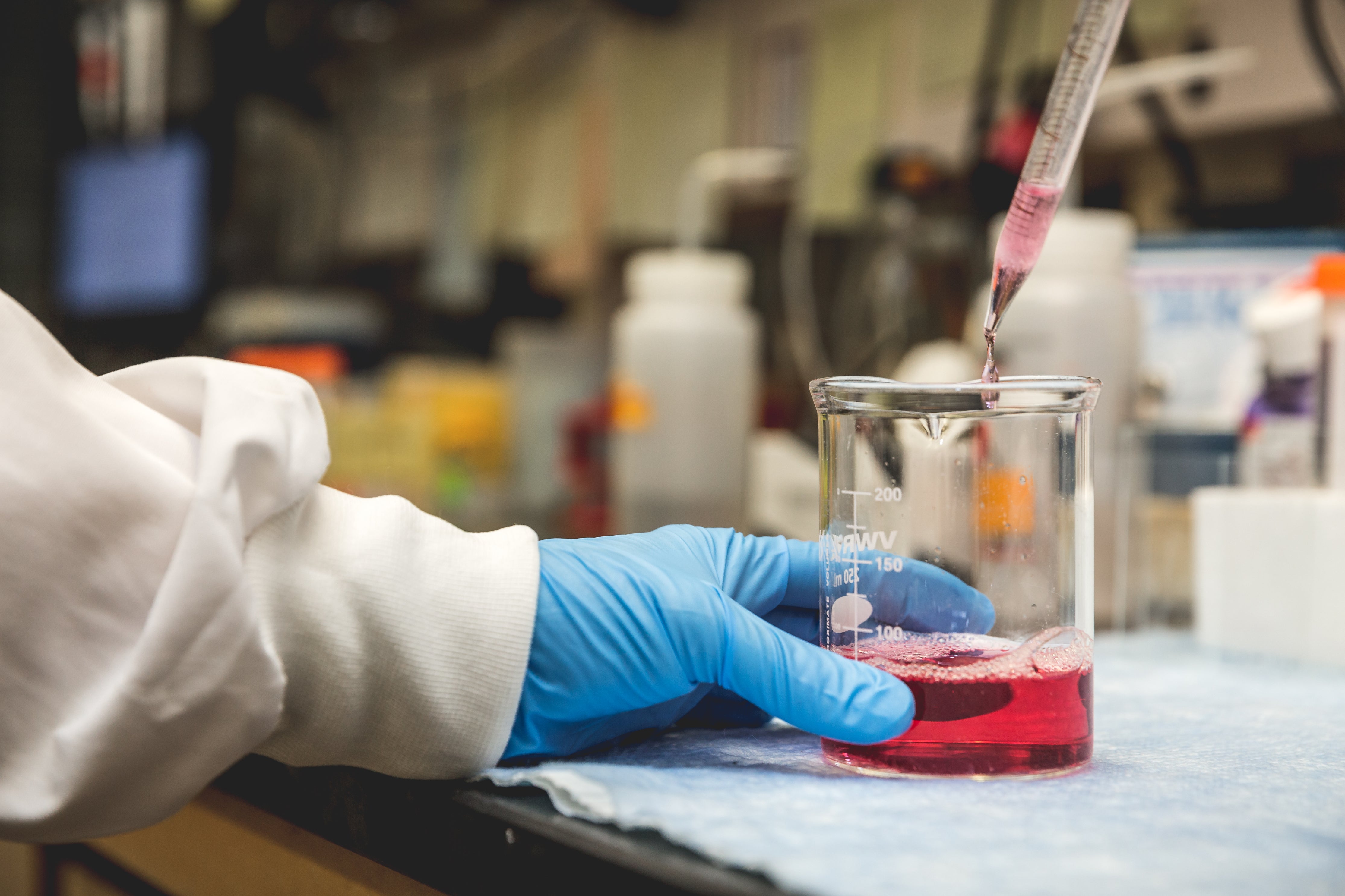 Gloved hand pipetting a red sample into a beaker in a lab