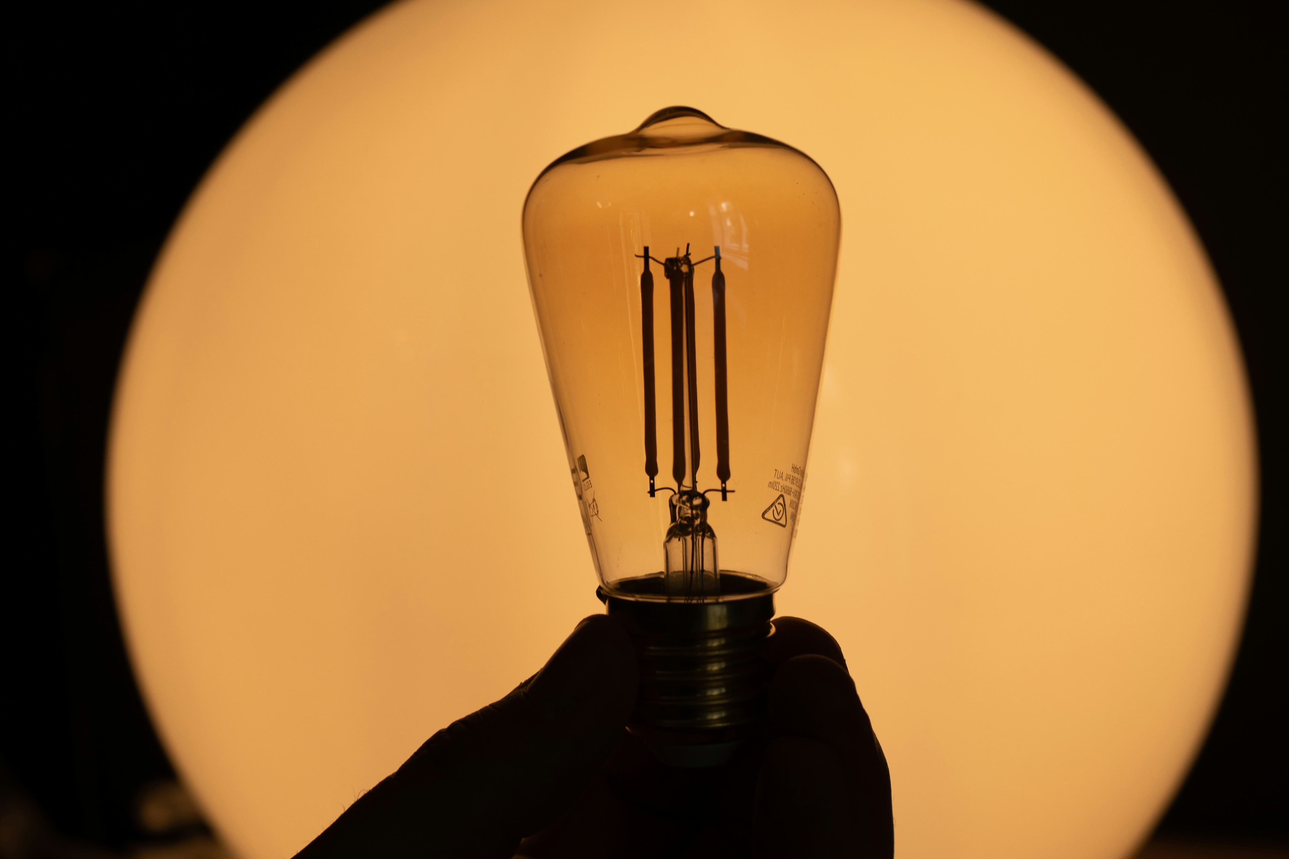 A hand holding a glowing filament light bulb against a warm circle of light