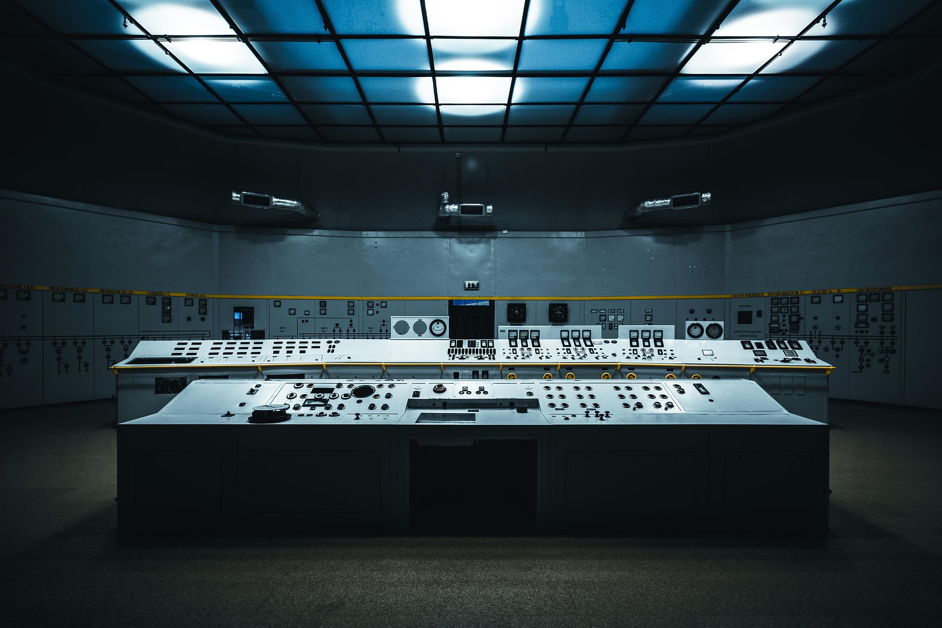 A vintage power station control room with long consoles of dials and switches under a grid of cool ceiling lights.