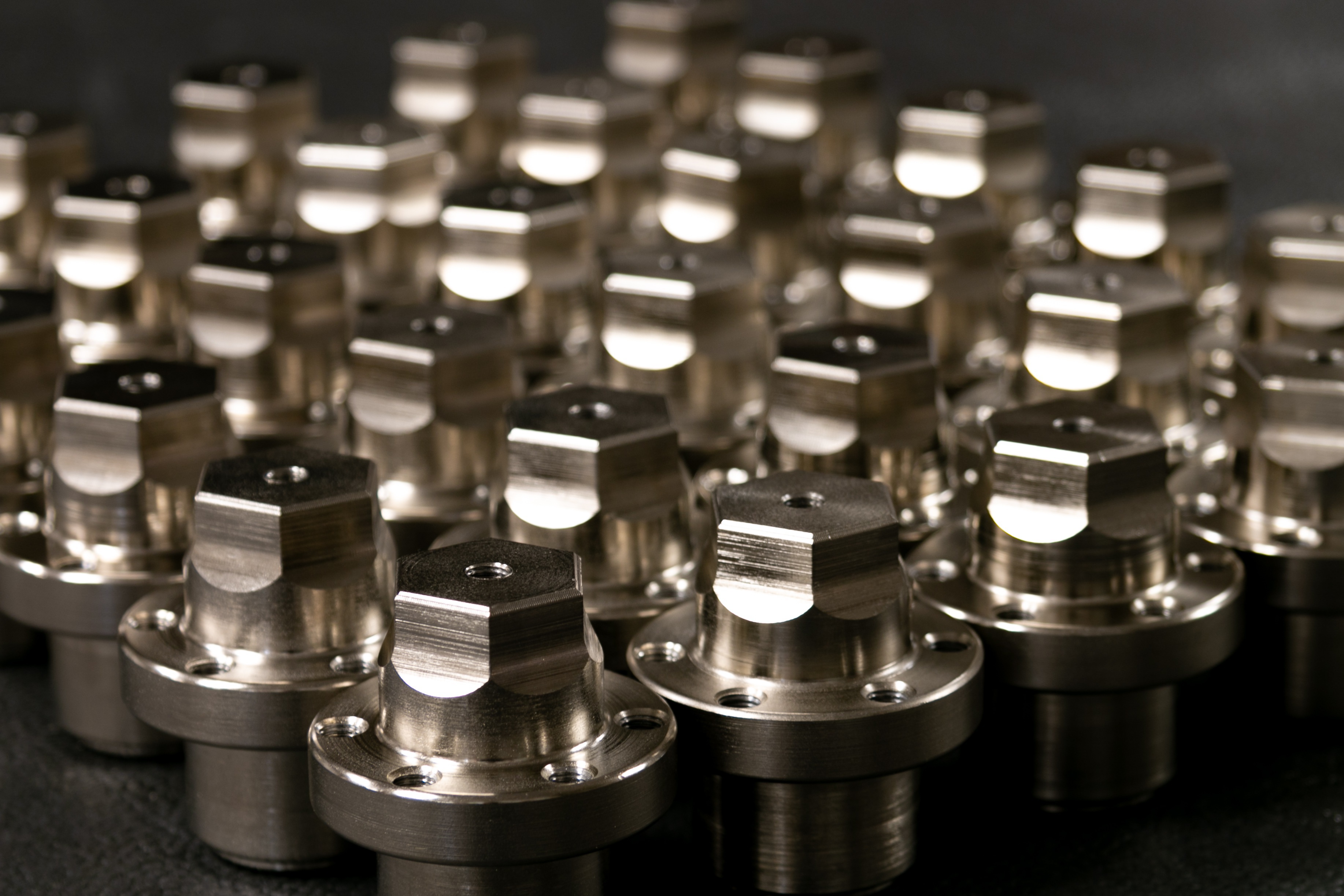 Rows of identical polished CNC-machined metal parts with hexagonal heads and flanges on a dark surface.